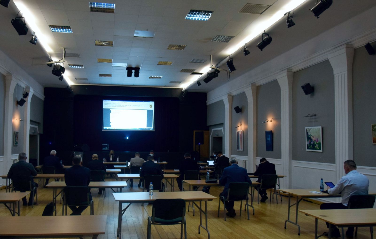Powiększ zdjęcie. Radni siedzą tyłem przy ławkach na dużej sali
