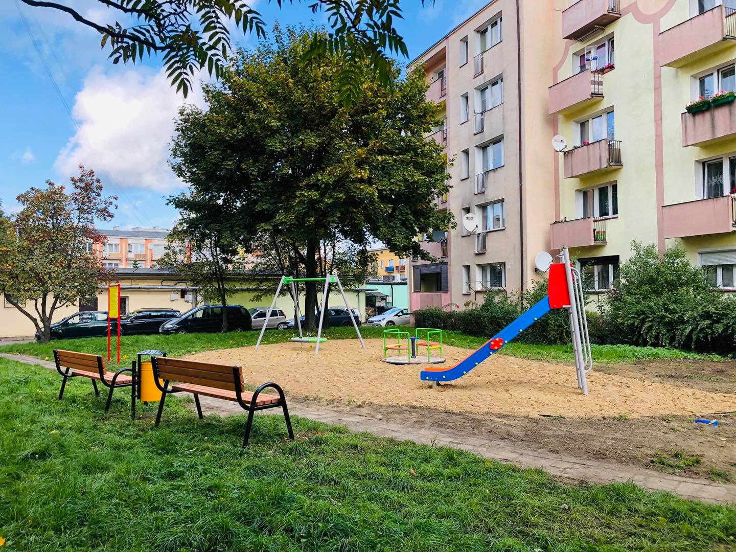 Plac zabaw: karuzela, zjeżdżalnia, karuzela, tablica, ławki, niebo, drzewo, blok, samochody