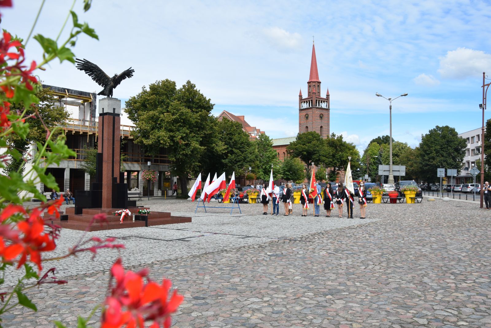 Powiększ zdjęcie. Widok na plac, pomnik, flagi Polski i poczty sztandarowe ze szkół