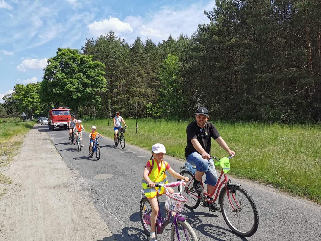Powiększ zdjęcie. Osoba dorosła i dziecko jadą rowerami, drzewa, niebo
