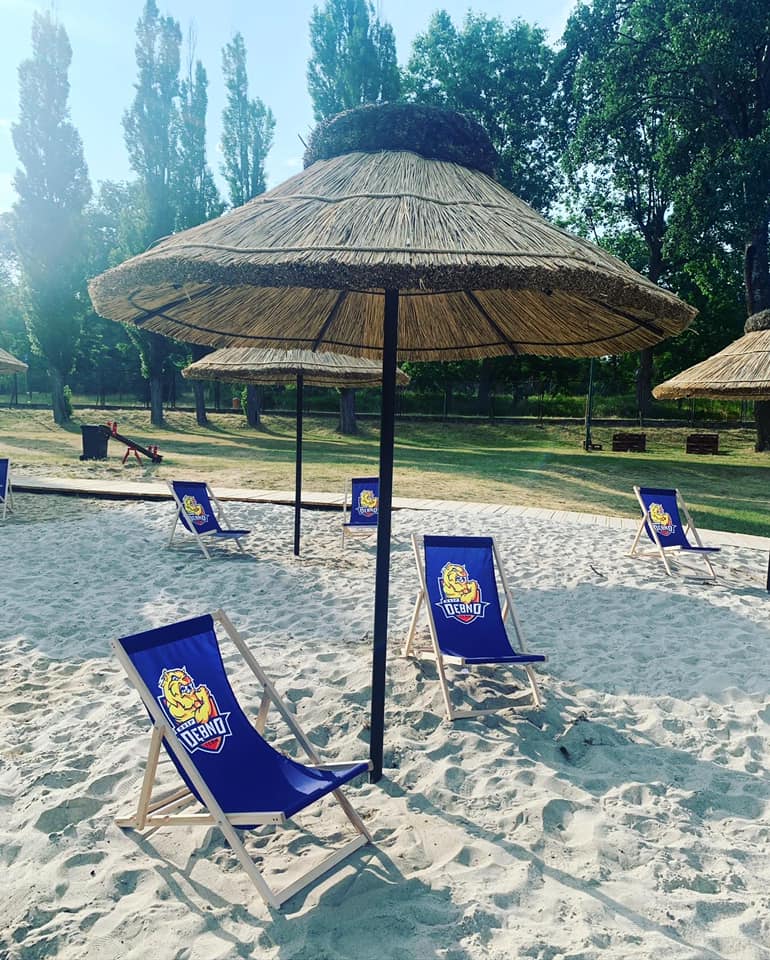 Powiększ zdjęcie. Parasol ze strzechy, leżaki na piasku, drzewa, niebo 