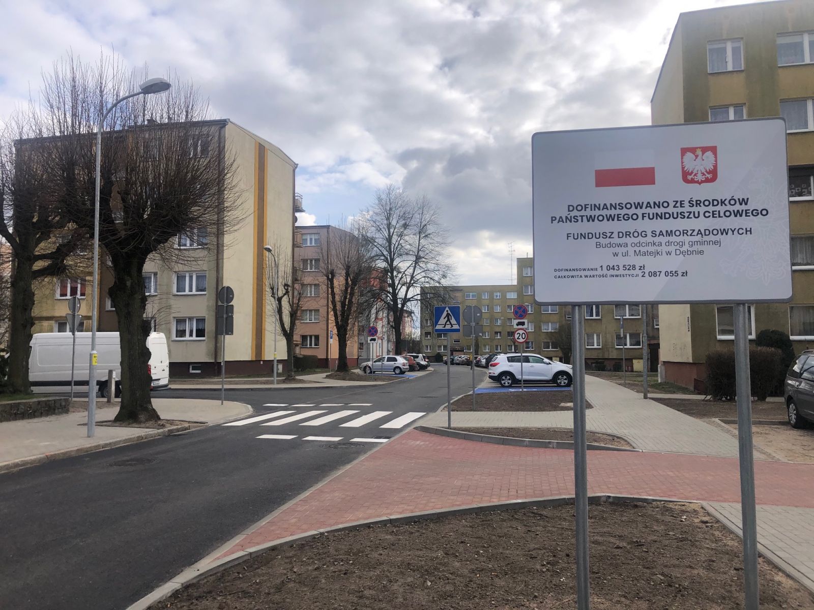Tablica z informacją o dofinansowaniu, lampa, niebo, bloki, drzewa, jezdnia, samochody