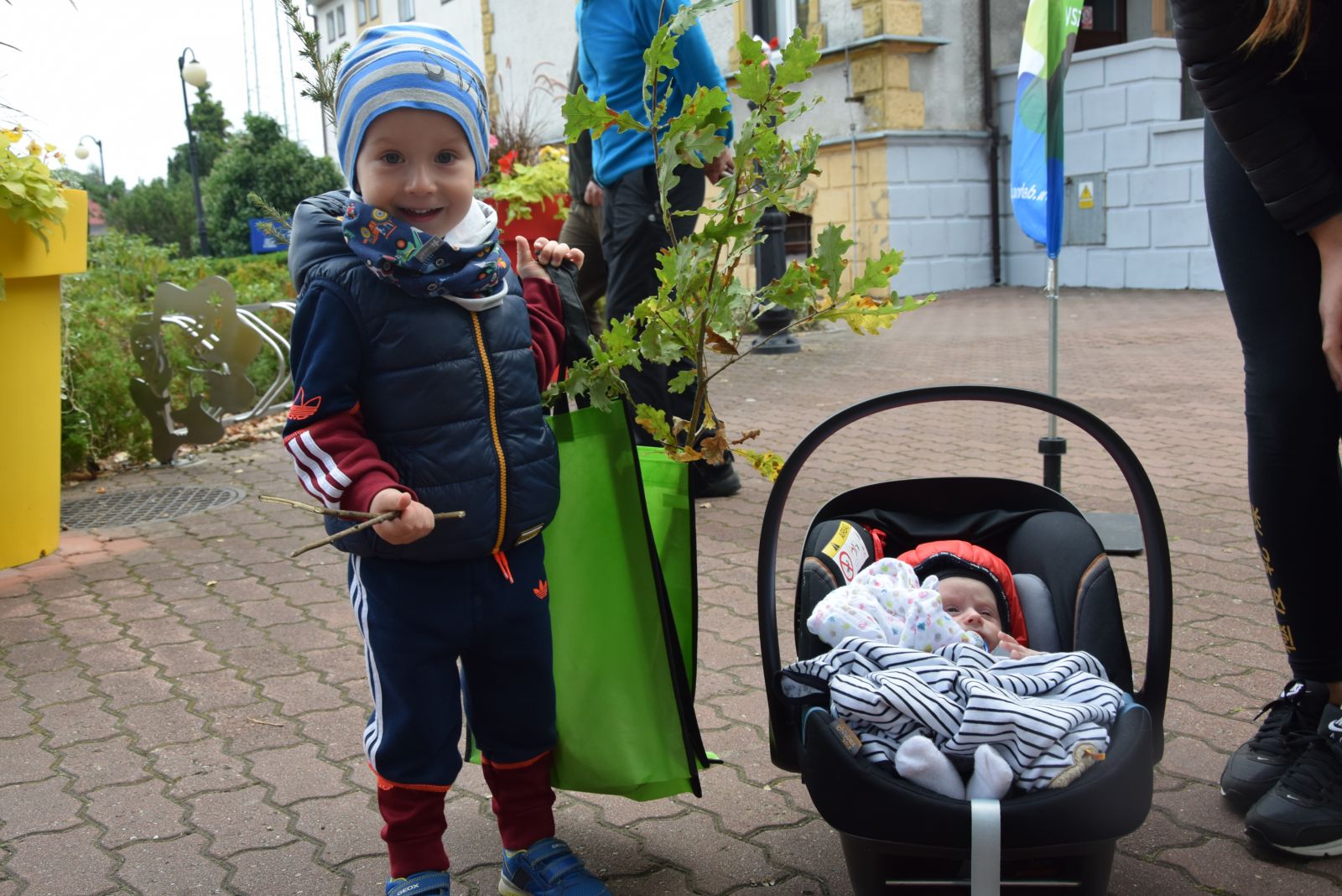Chłopczyk z drzewkiem, obok dziecko w nosidełku