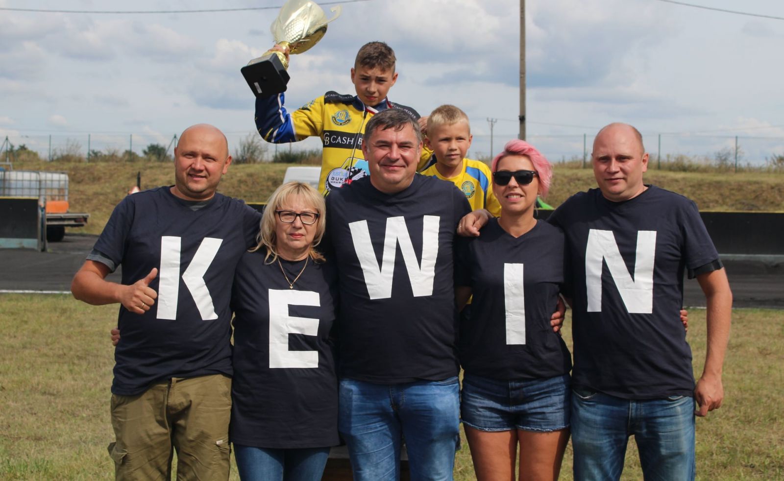 Powiększ zdjęcie. Kewin Nycz w ręku trzyma puchar. Przed nim ludzie w koszulkach, napis KEWIN
