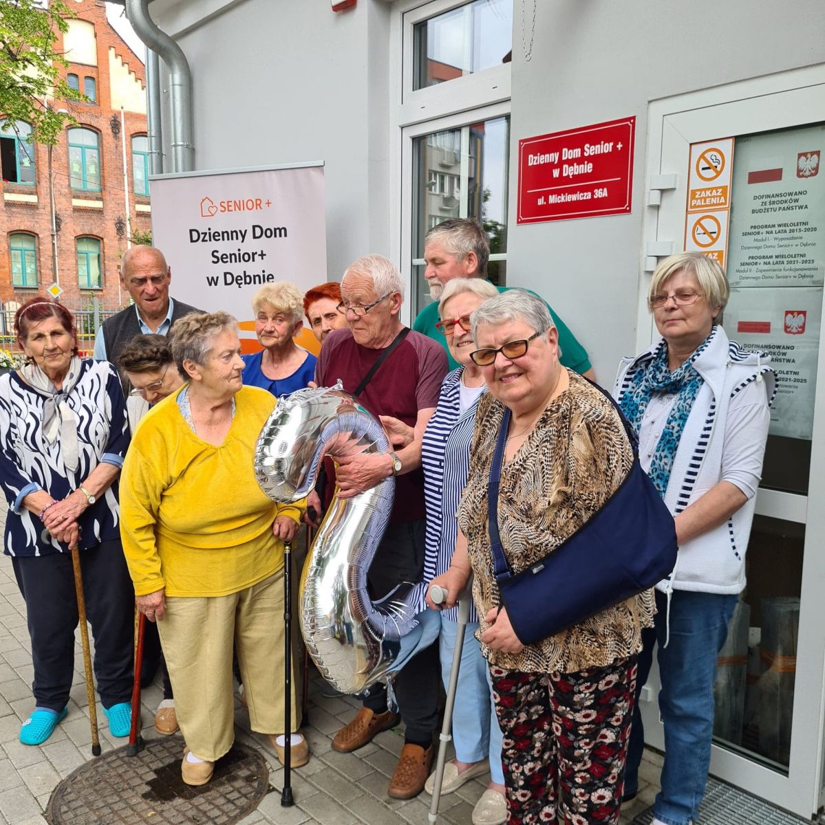 Powiększ zdjęcie. Seniorzy przed wejściem do budynku, w ręku trzymają balon z numerem 2