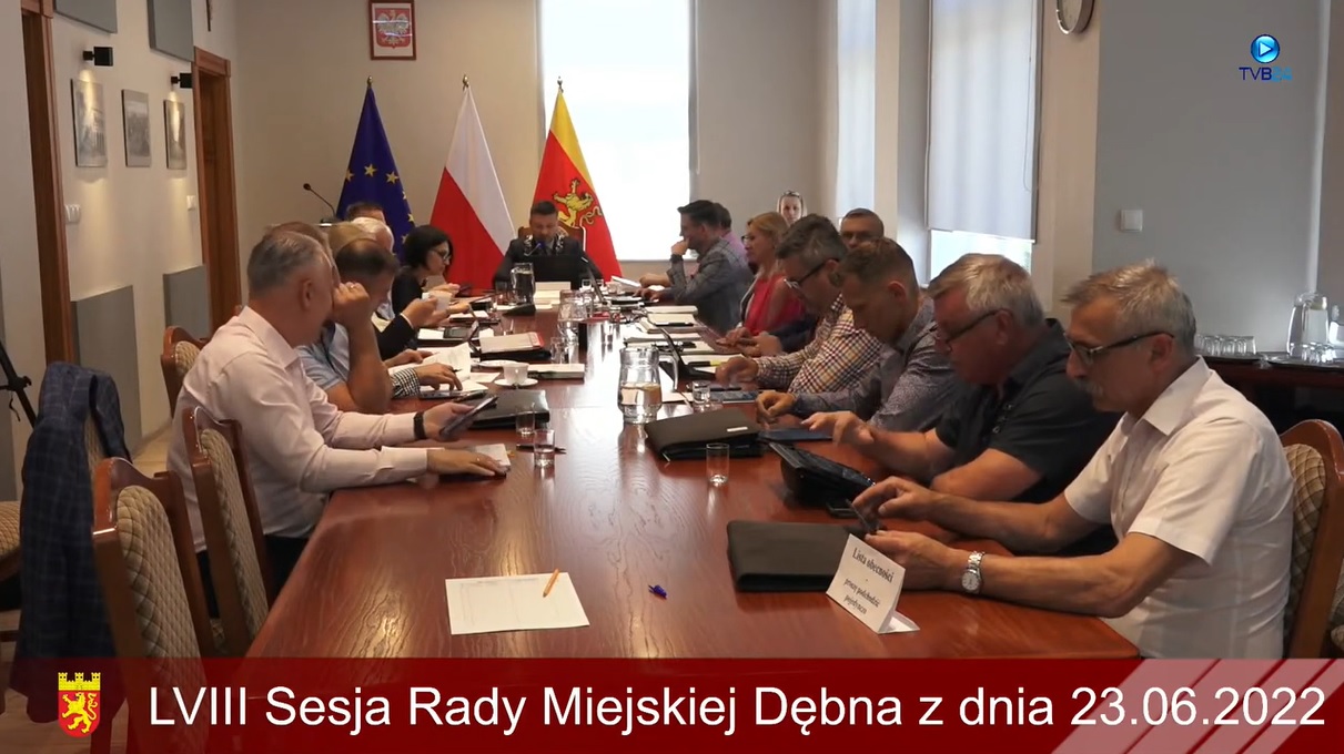 Powiększ zdjęcie. Radni siedzą przy stole w sali posiedzeń. w tle flagi 