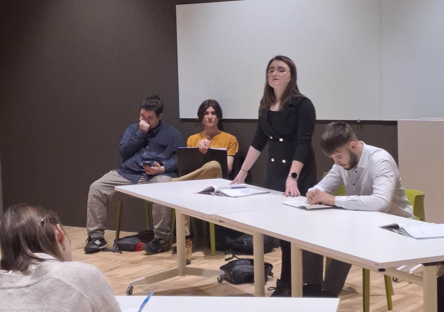 Powiększ zdjęcie. Dziewczyna stoi przy stole, coś mówi