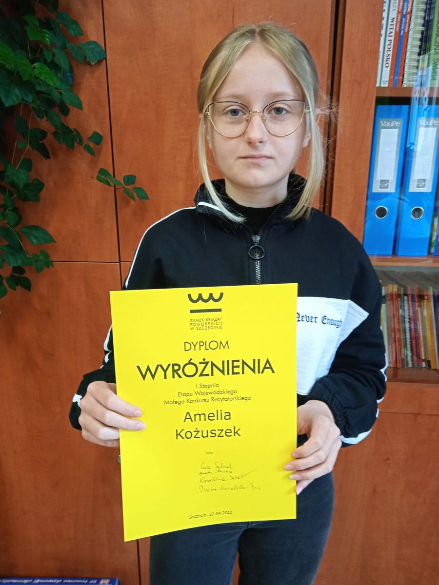 Powieksz zdjęcie. Amelia w pomieszczeniu trzyma w ręku żółty dyplom