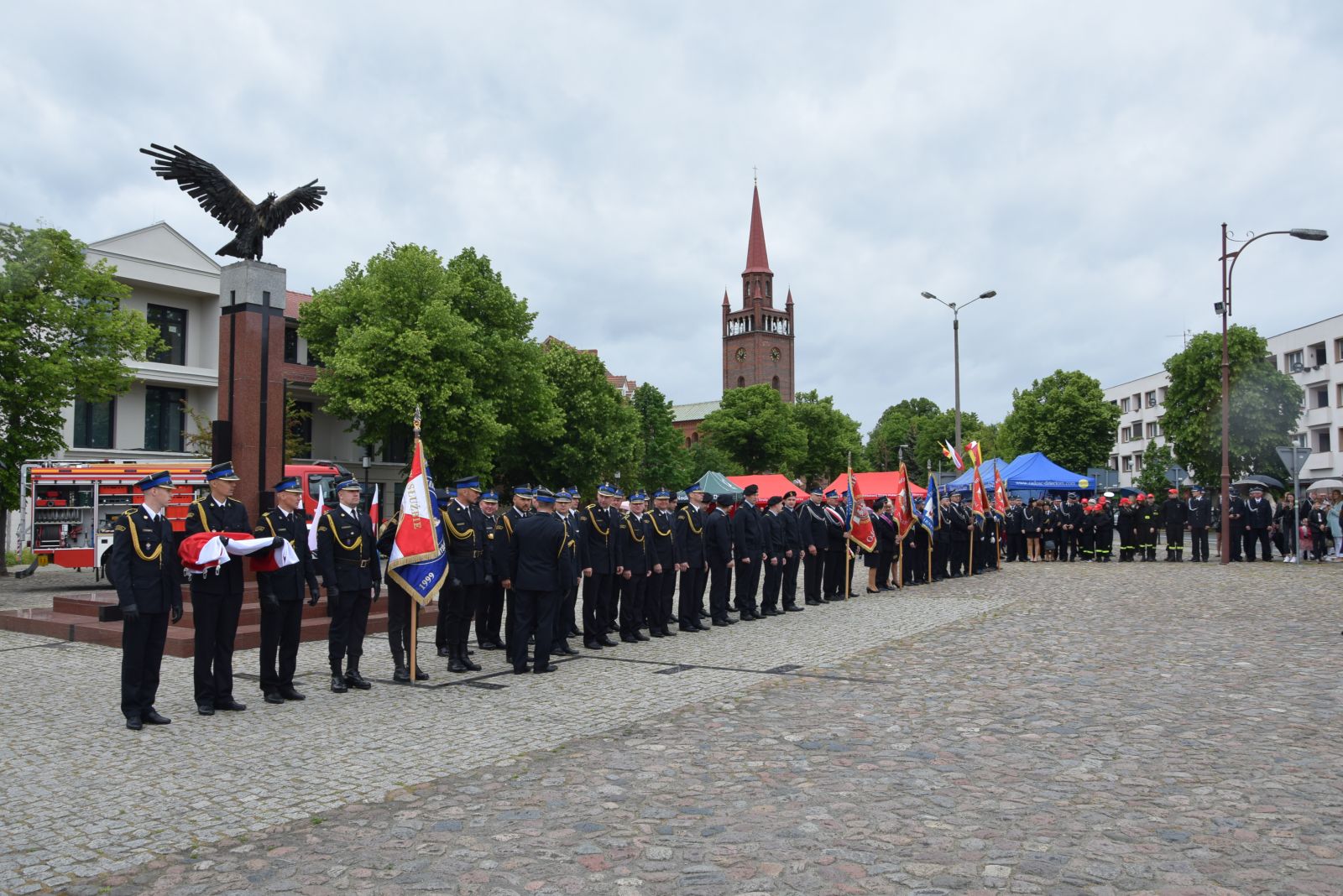 Powiększ zdjęcie. Strażacy stoją w szeregu na placu