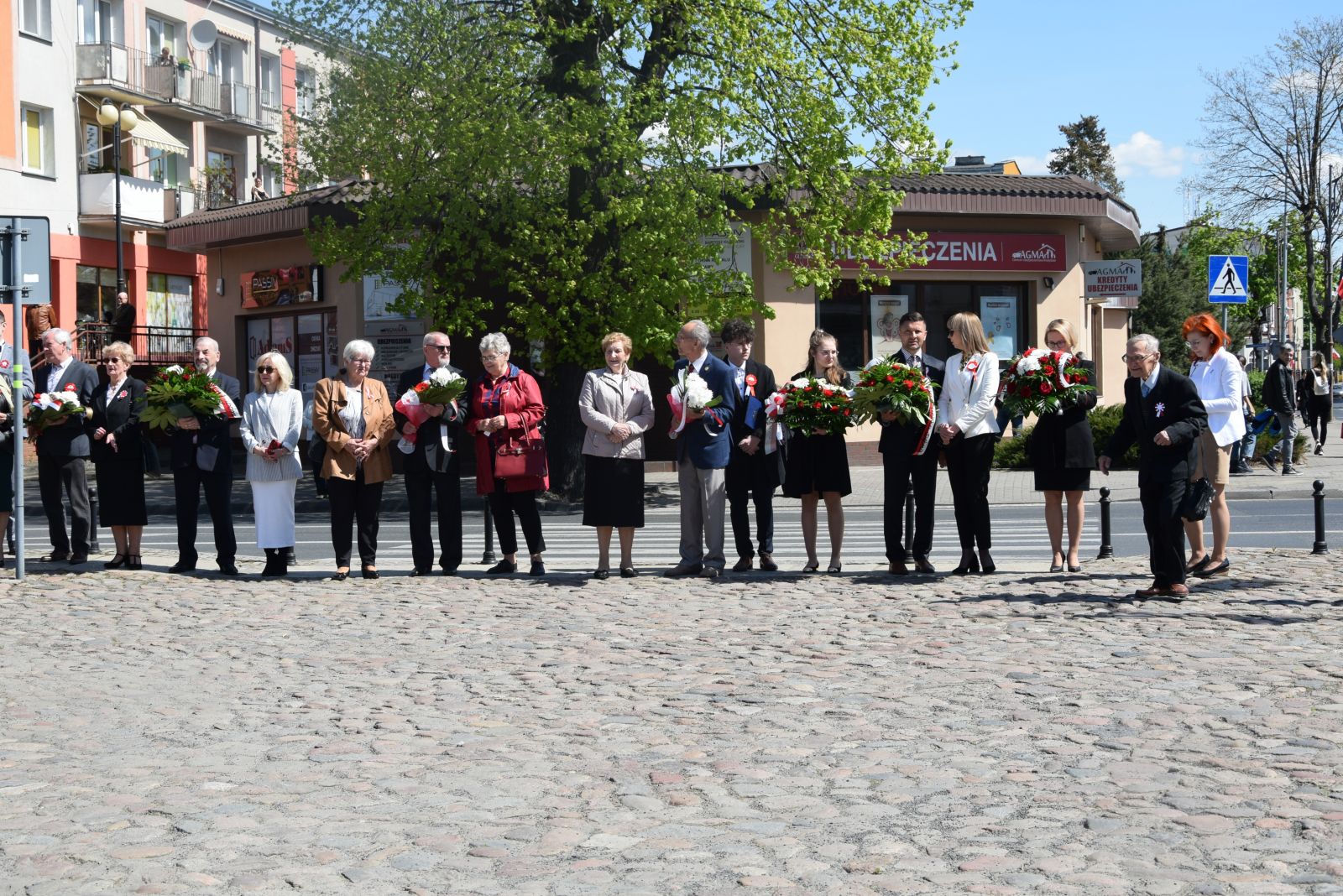 Powiększ zdjęcie. Uczestnicy uroczystości stoją w rzędzie, trzymają kwiaty  