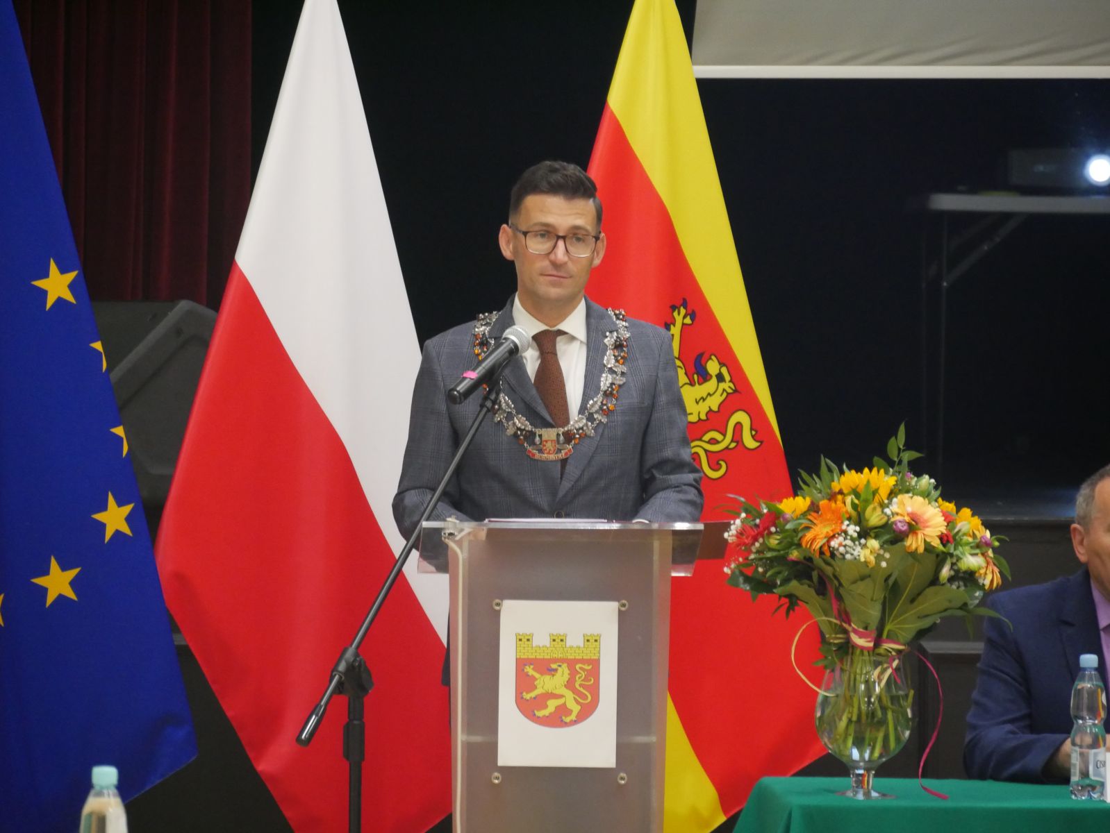 Powieksz zdjęcie. Burmistrz przemawia, w tle flagi UE, Polski, Dębna
