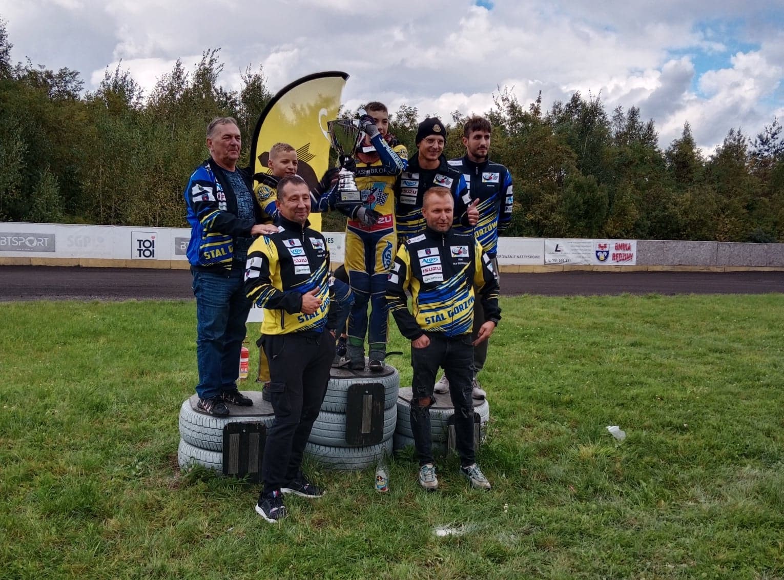 Kewin Nycz stoi na najwyższym podium, trzyma puchar, obok kolega, wokół trenerzy 