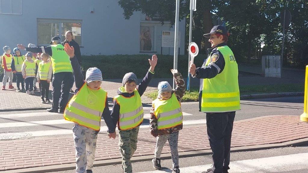 Dzieci w kamizelkach odblaskowych przechodzą przez pasy z wysepką przy eskorcie  