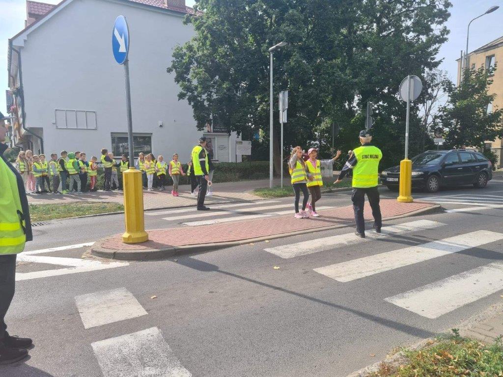 Dzieci przechodzą przez przejście z wysepką, przy eskorcie strażnika 