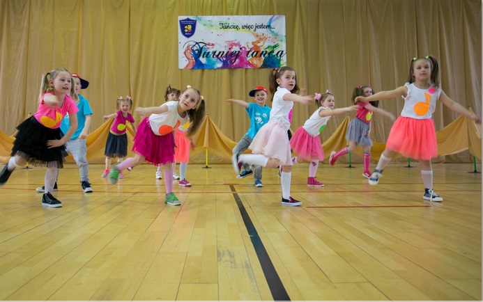 Powiększ zdjęcie. Grupa dzieci, w kolorowych ubraniach tańczy. W pierwszej lini cztery dziewczynki