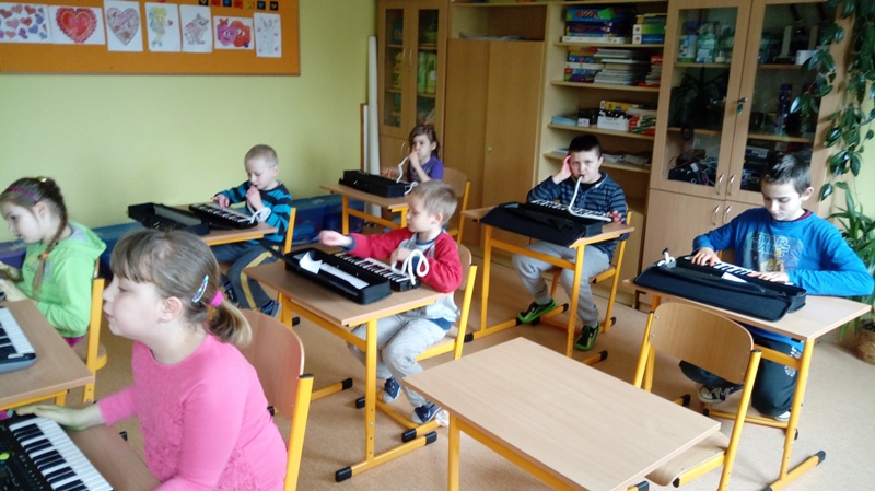 Powiększ zdjęcie. Uczniowie siedzą przy ławkach szkolnych i grają na małych keyboardach