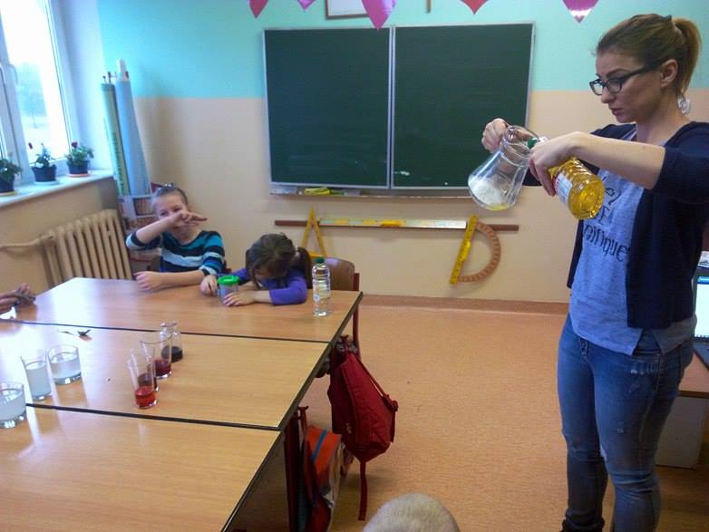 Nauczyciel do dzbanka nalewa olej. W tle w ławce siedzą dwie dziewczynki