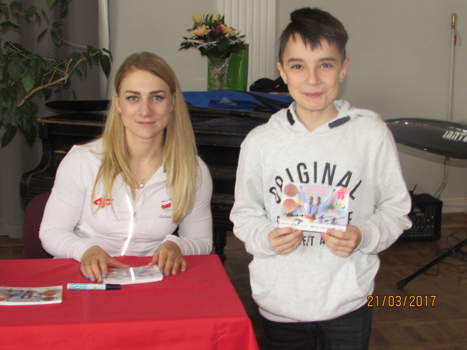 Powiększ zdjęcie. Karolina Naja siedzi przy stole, obok stoi uczeń, trzyma w ręku zdjęcie z autografem