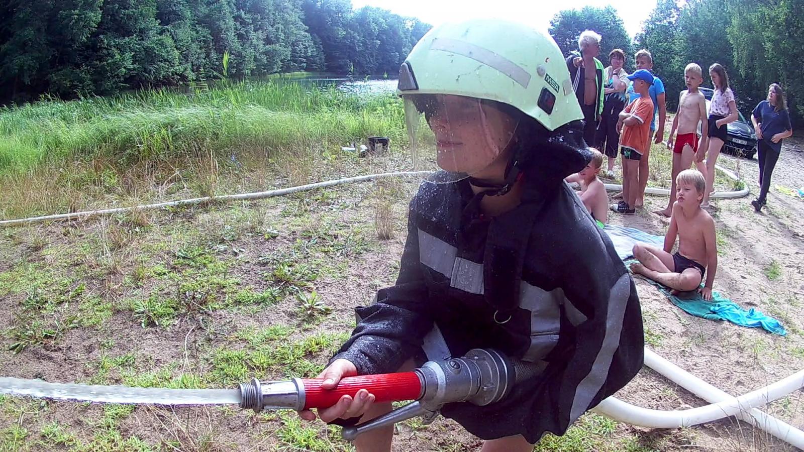 Zdjęcie dziewczynki w stroju strażaka z wężem gaśniczym.