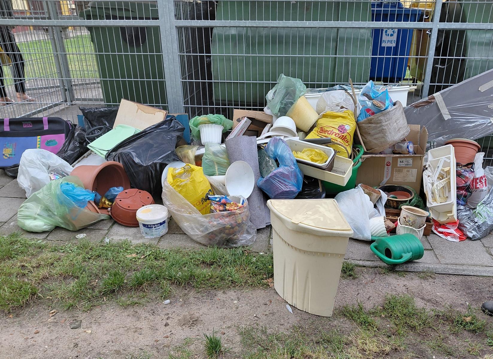 Śmieci porzucone przy boksie z odpadami, ogrodzenie metalowe, za nim zielone kubły na odpady