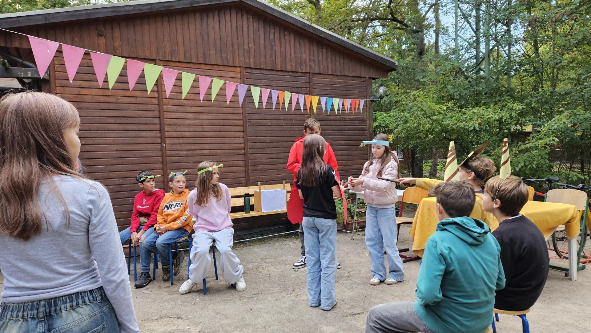 Dzieci na placu siedzą na krzesełkach, inne stoją, nad nimi wisi kolorowa gurlanda z trójkątów, w tle drewniana ściana budynku