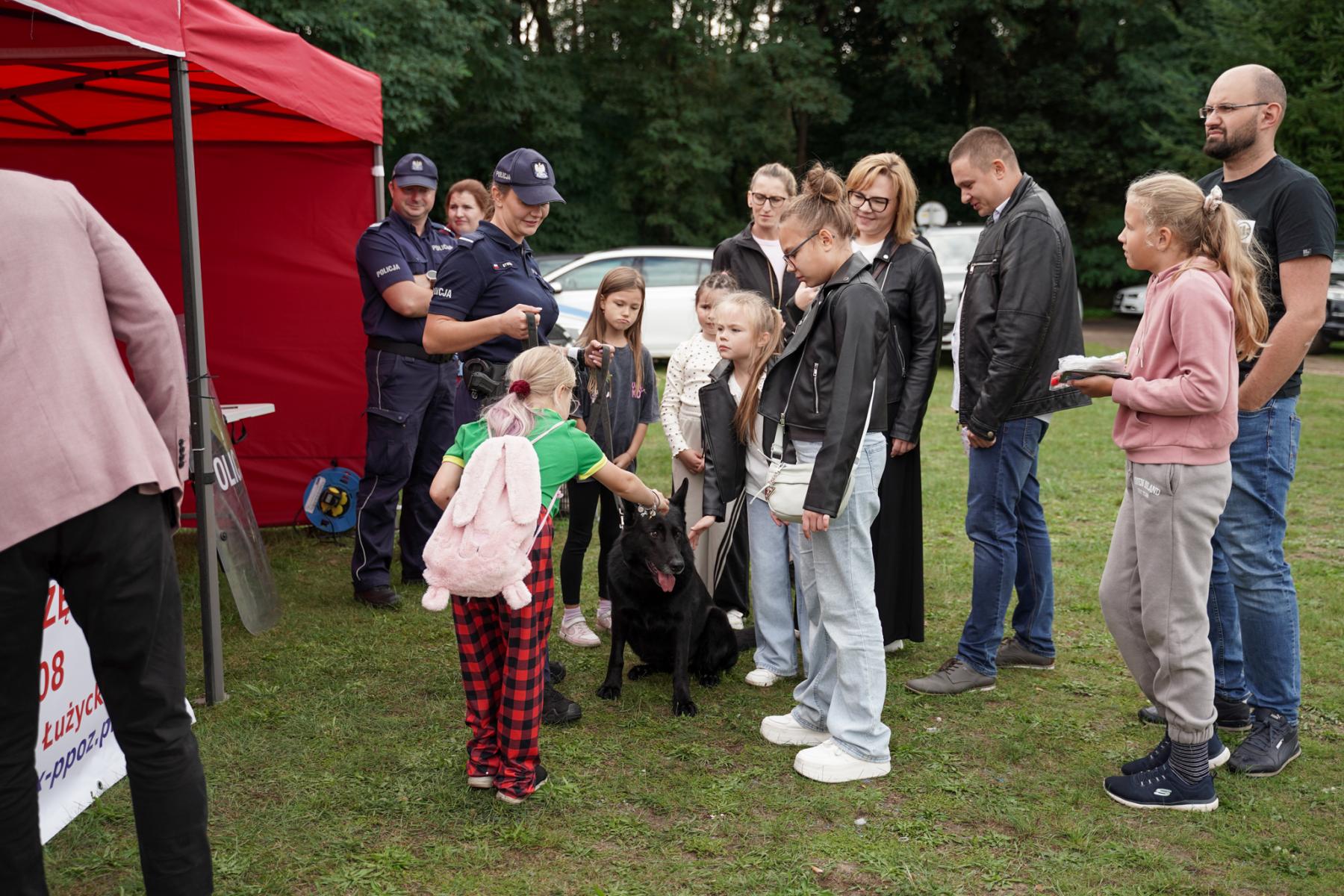 Ludzie stoją przer czerwonym namiotem, Policjantka trzyma na smyczy czarnego psa, dziewczyna głaszcze go