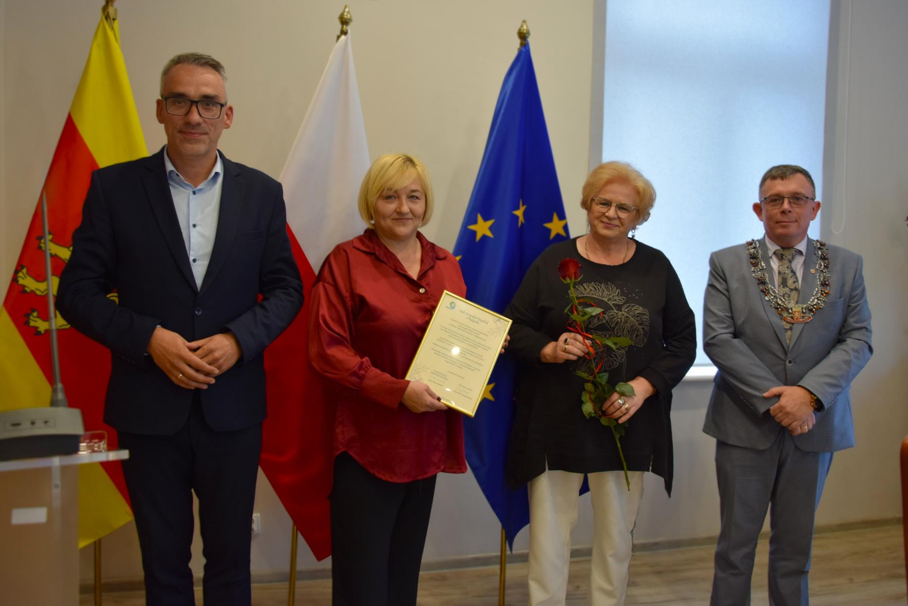 Od lewej stoją w sali: burmistrz, dwie kobiety, przewodniczący, w tle flagi: gminna żółto - czerwona, polski biało-czerwona, UE niebieska