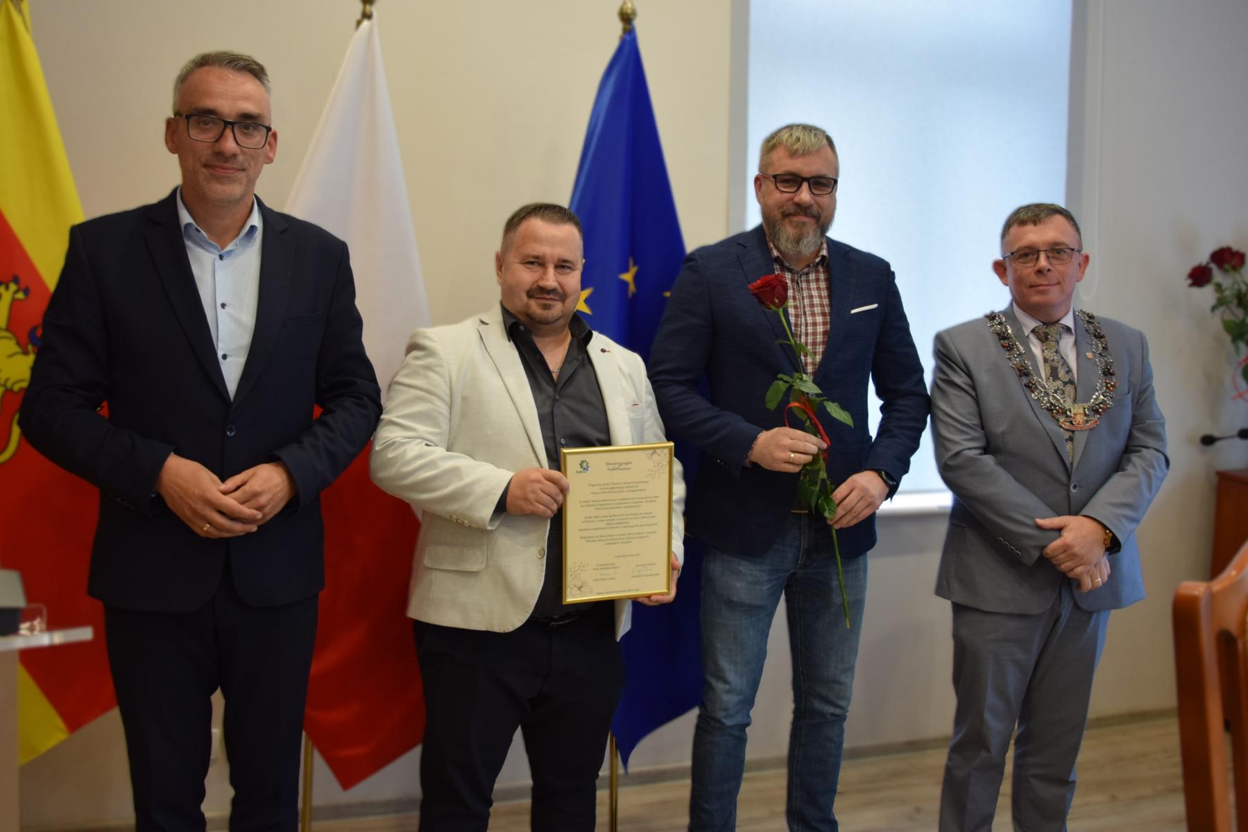 Od lewej stoją w sali: burmistrz, dwóch mężczyzn, przewodniczący, w tle flagi: gminna żółto - czerwona, polski biało-czerwona, UE niebieska