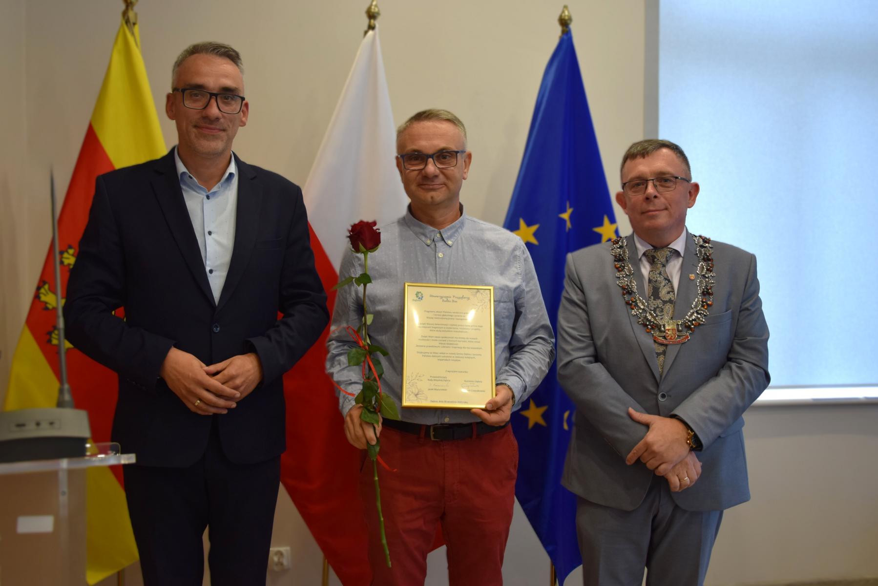Od lewej stoją w sali: burmistrz, mężczyzna , przewodniczący, w tle flagi: gminna żółto - czerwona, polski biało-czerwona, UE niebieska