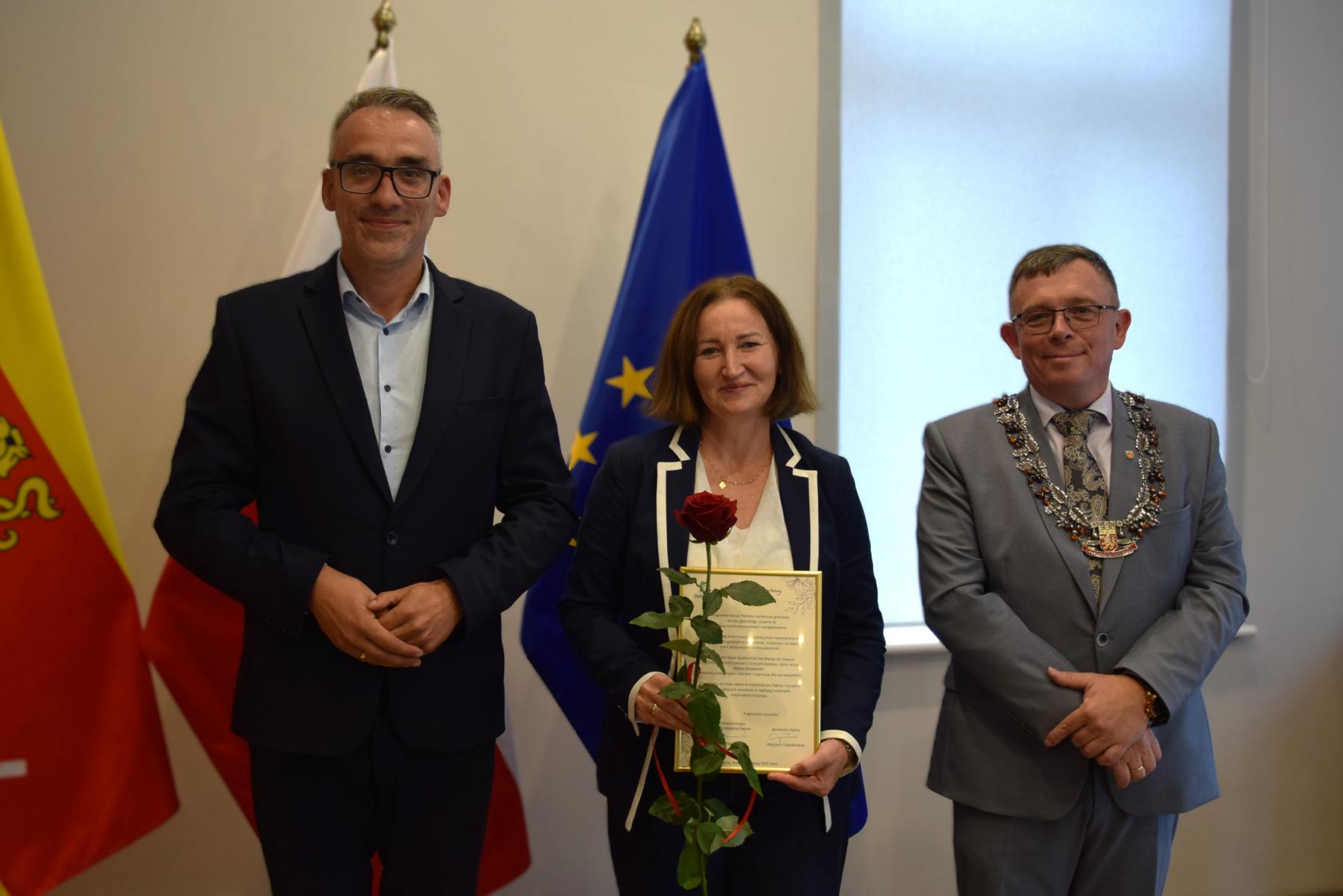 Od lewej stoją w sali: burmistrz, kobieta, przewodniczący, w tle flagi: gminna żółto - czerwona, polski biało-czerwona, UE niebieska