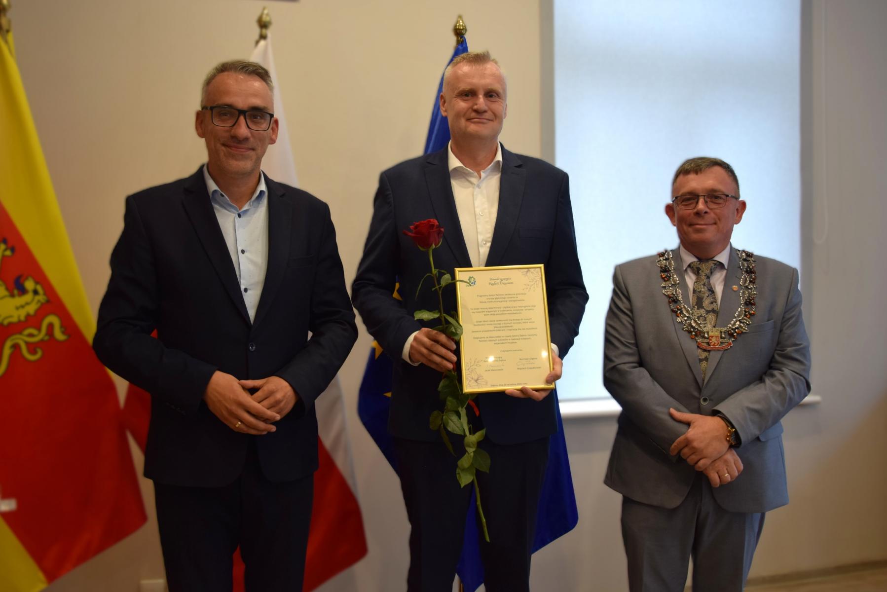 Od lewej stoją w sali: burmistrz, mężczyzna , przewodniczący, w tle flagi: gminna żółto - czerwona, polski biało-czerwona, UE niebieska