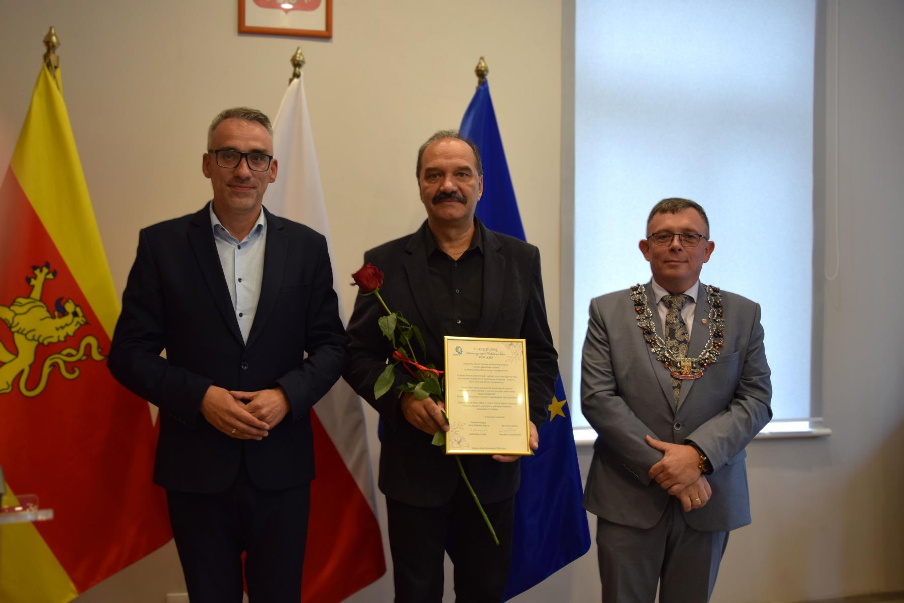 Od lewej stoją w sali: burmistrz, mężczyzna, przewodniczący, w tle flagi: gminna żółto - czerwona, polski biało-czerwona, UE niebieska