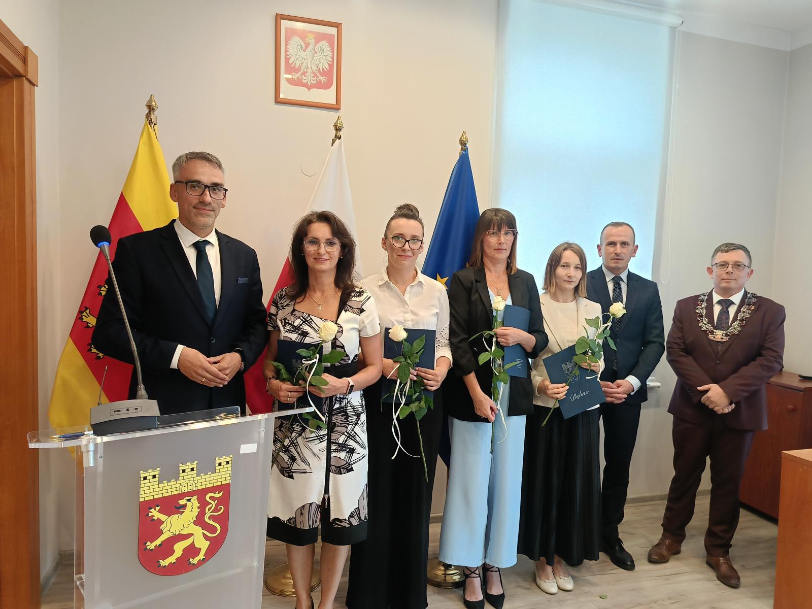 Od lewej stoją w sali: mężczyzna (burmistrz Dębna), cztery panioe elegancko ubrane, mężczyzna (wicemarsząłkre województwa). Każda z Pań trzyma granatową teczkę i białą różę. 