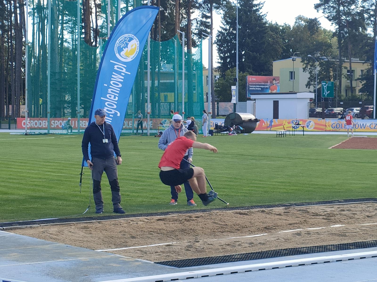 Karol Radziukieiwcz skacze w dal. obok na trawie stoi dwóch mężczyzn