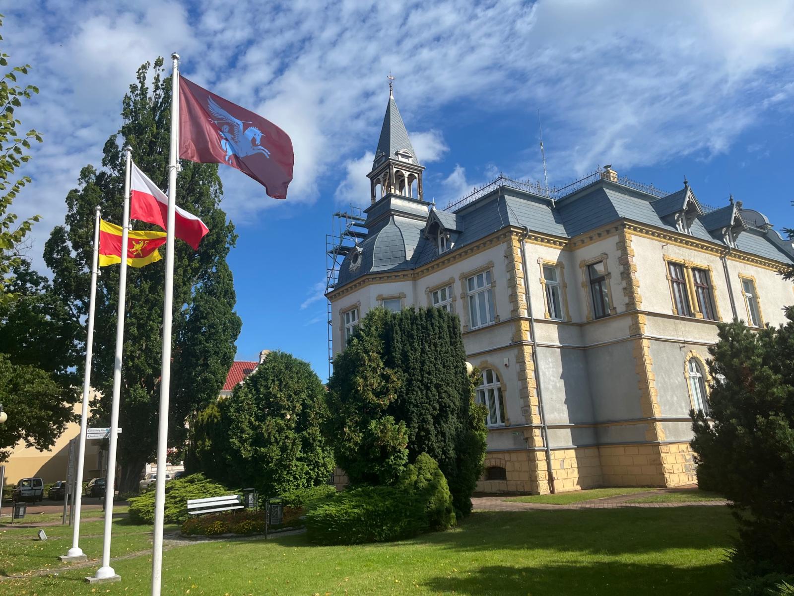Flagi powiewające na masztach, w tle budynek urzędu, niebieskie niebo, drzewa