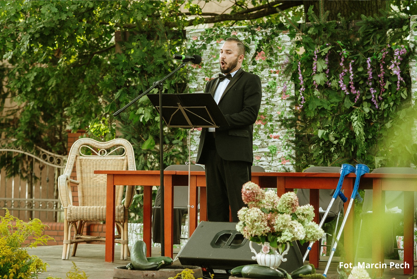 Śpiewak, elegancko ubrany, stoi na podeście w zielonej scenerii, 