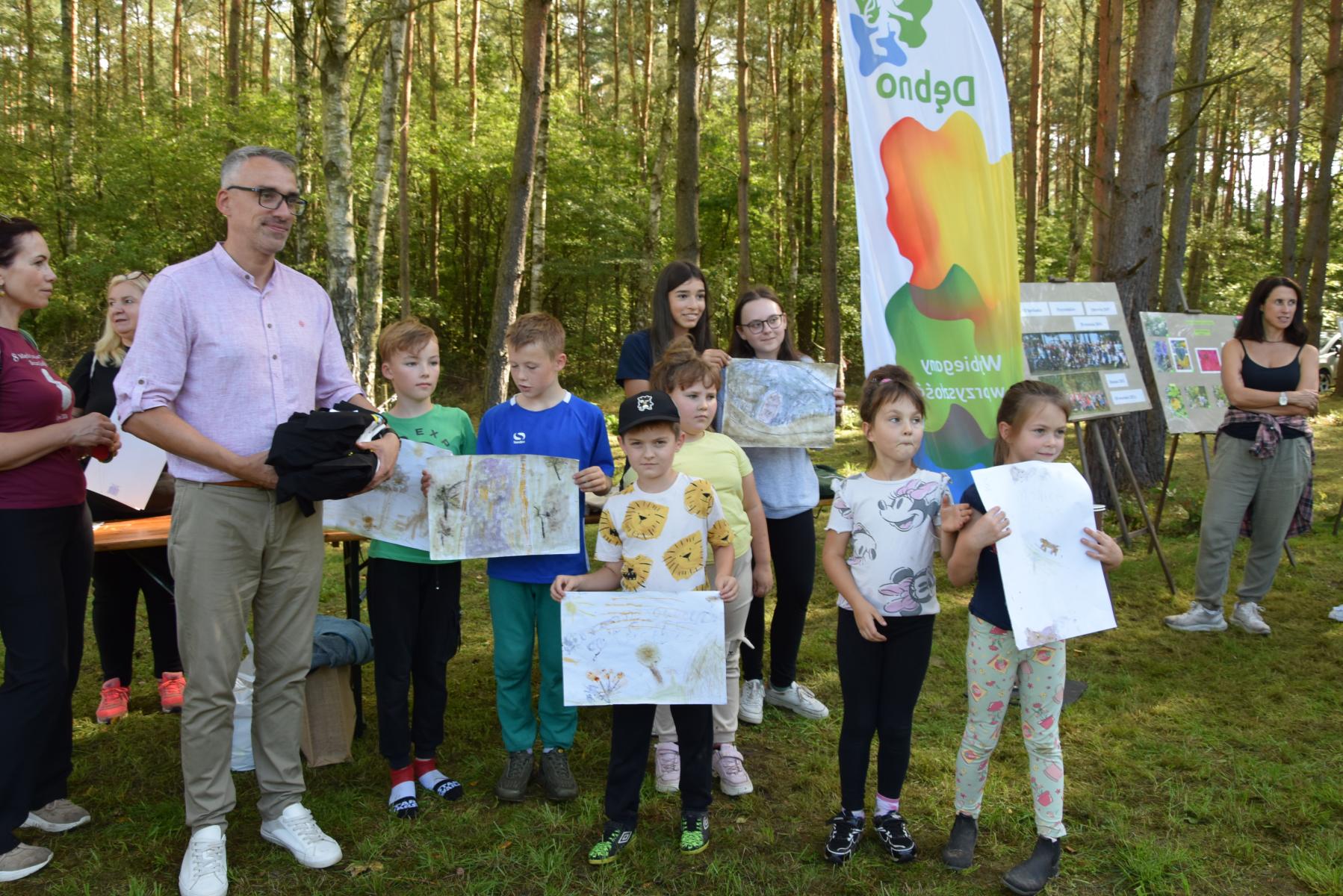 Grupa uczniów stoi z pracami plastycznymi, od lewej stoi burmistrz Dębna, w tle drzewa  