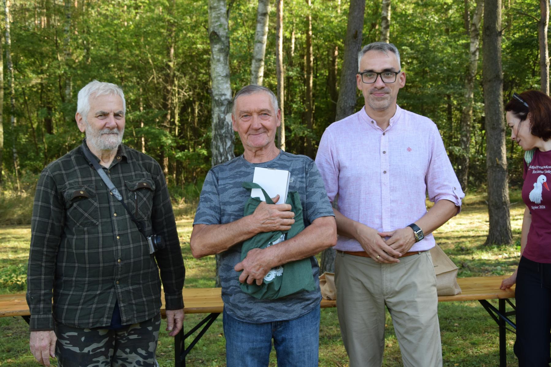 Od lewej stoją: Marek Schiller, dorosły mężczyzna ściska worek w ręku, Wojciech Czepułkowski, w tle las i zieleń
