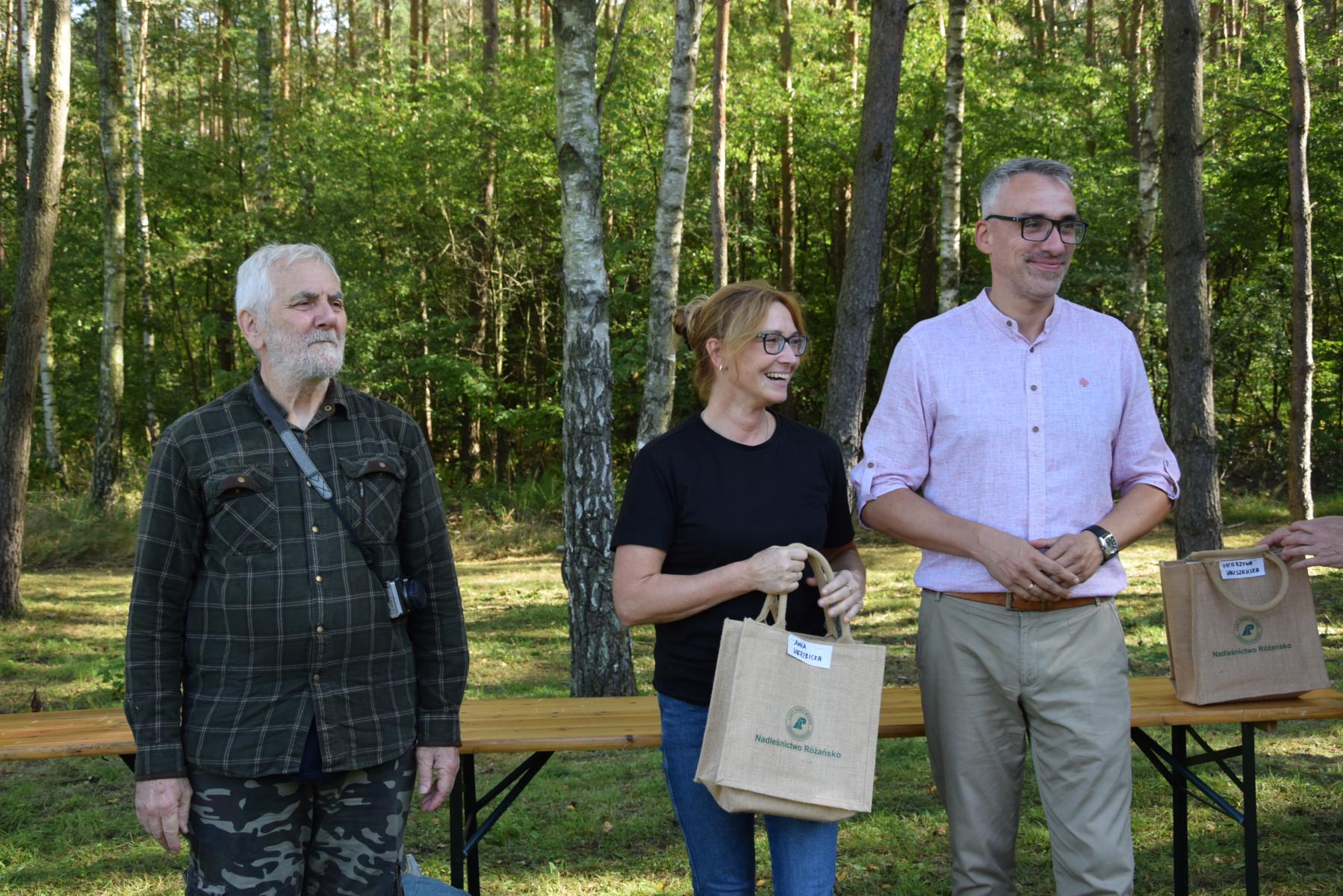 Od lewej stoją: Marek Schiller, kobieta uśmiechnięta z torbą w ręku, Wojciech Czepułkowski, w tle las i zieleń