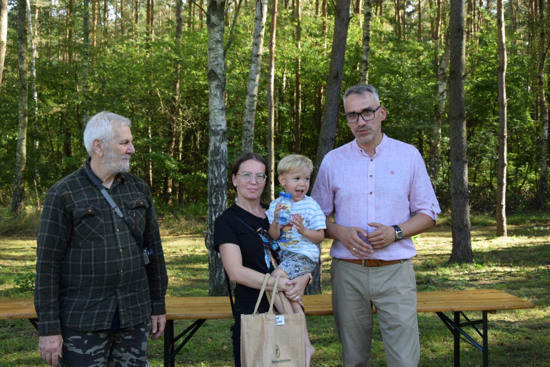 Od lewej stoją: Marek Schiller, kobieta z dzieckiem (chłopcem) na ręku, Wojciech Czepułkowski, w tle las i zieleń