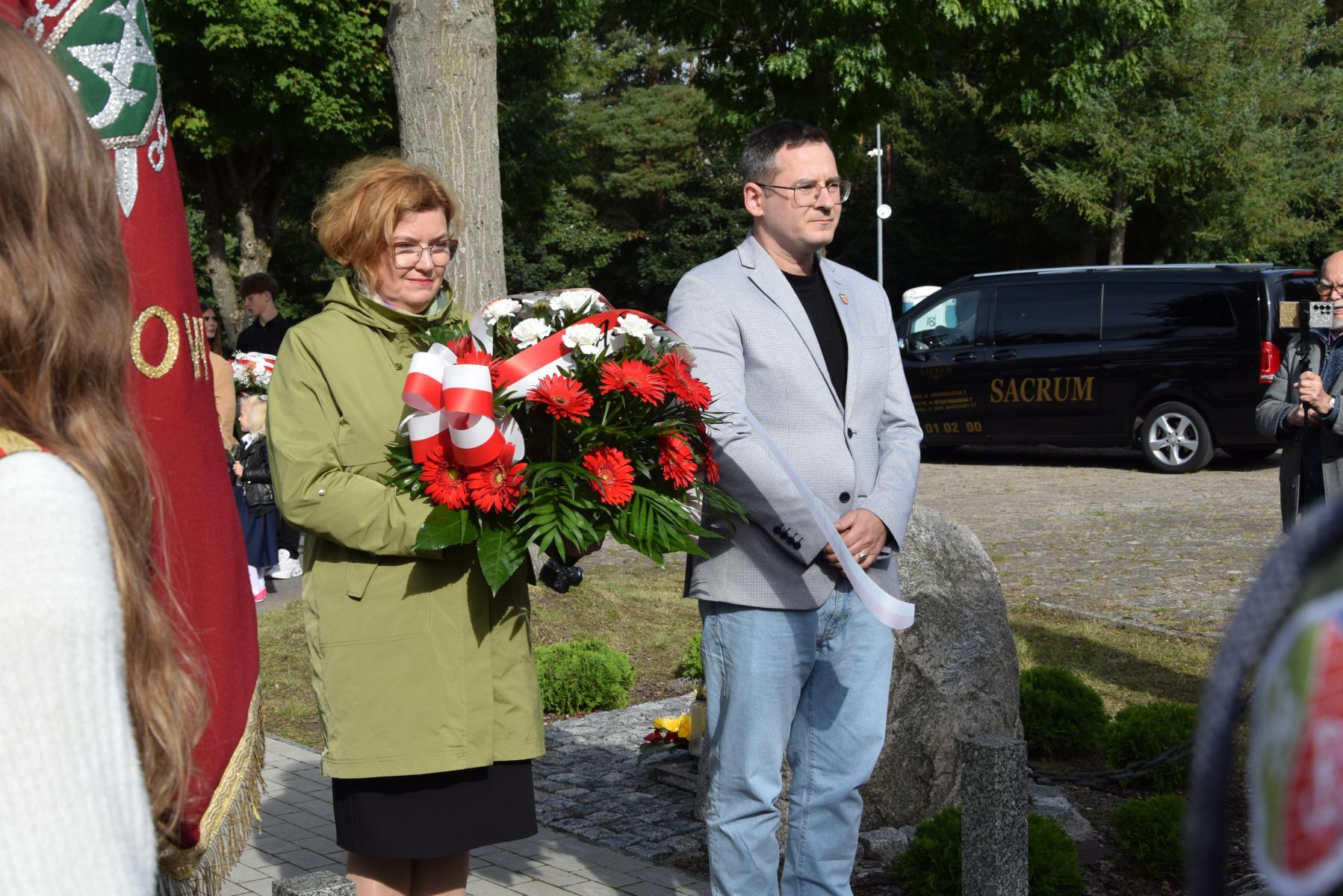 Delegacja samorządu: Kobieta z wieńcem biało-czerwonych kwiatów w dłoni, obok mężczyzna