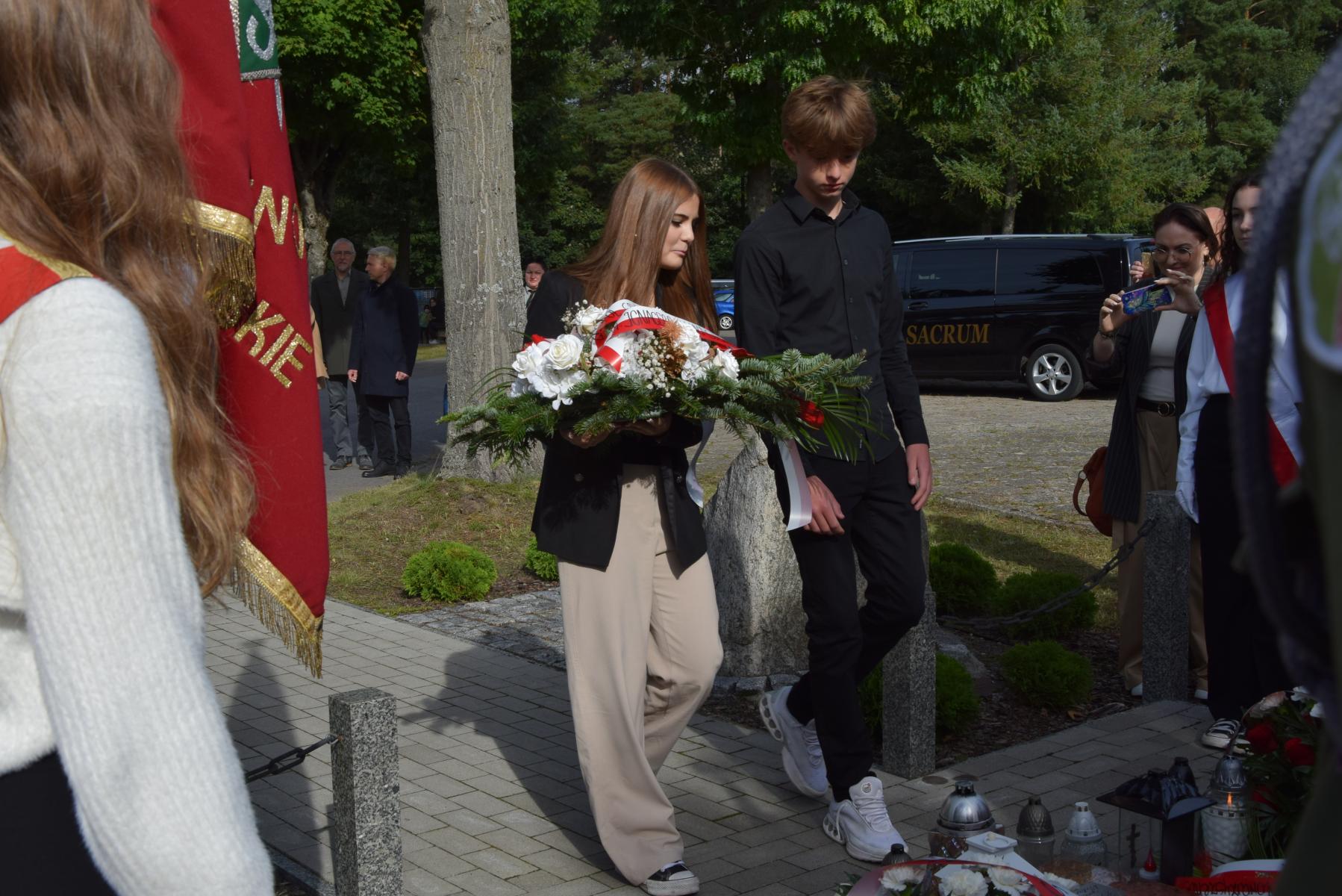 De3legacja młodzieży z Zespołu Szkół Stowarzyszenia Oświatowego; dziewczyna trzyma bukiet biało-czerwonych kwiatów, obolk stoi chłopak  