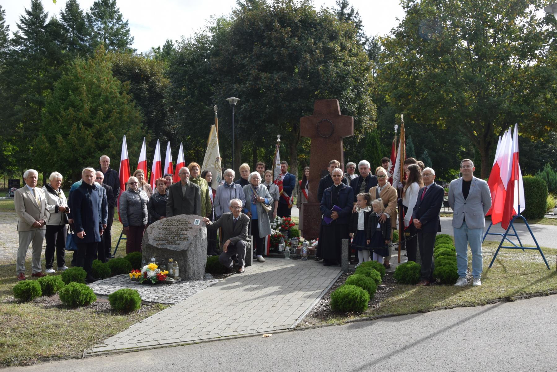 Grupa uczestników uroczystości stoi wokół, za i przed Pomnikiem Sybiraków, po bokach biało czerwone flagi 