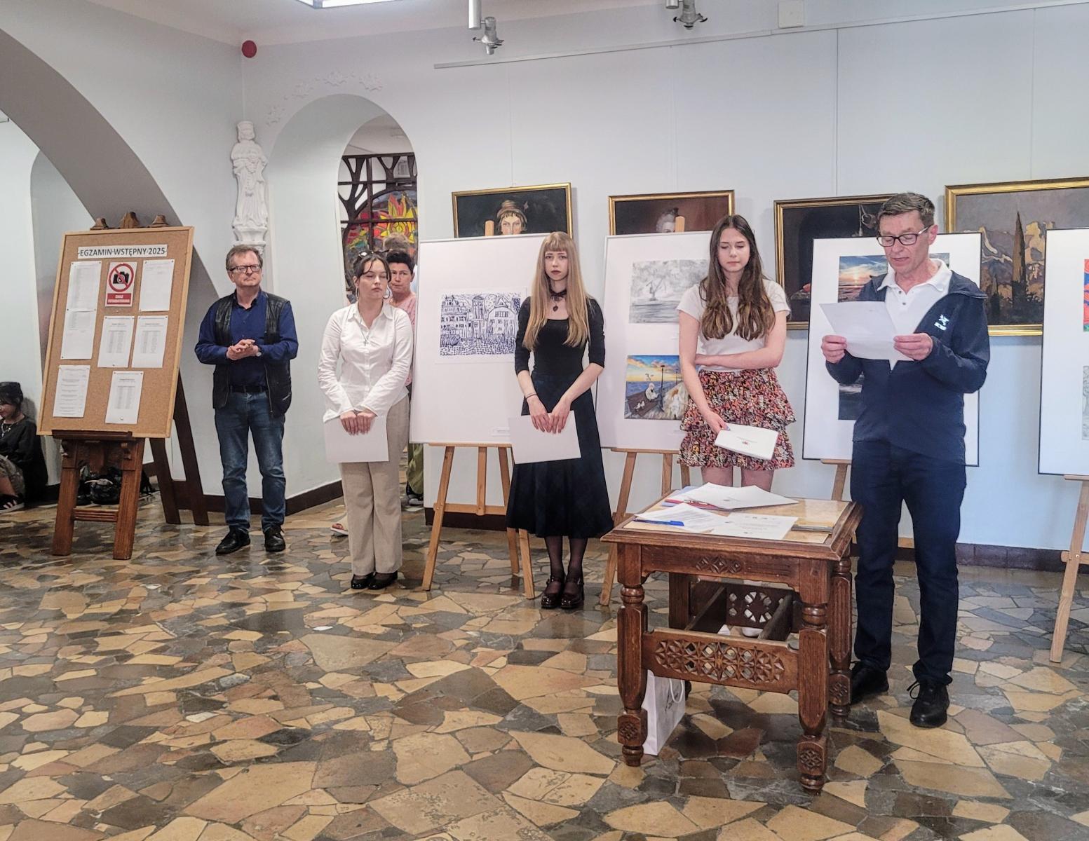 Pomieszczenie, Amelia ubrana w czarną sukienkę stoi cenralnie na zdjęciu. obok po prawej i lewej stronie inne dziewczyny. w prawym rogu  mężczyzna trzyma kartke i wygląda jakby czytał.