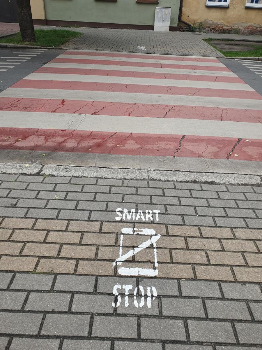 Przejście dla pieszych, na chodniku biały piktogram Smart STOP i przekreślony telefon
