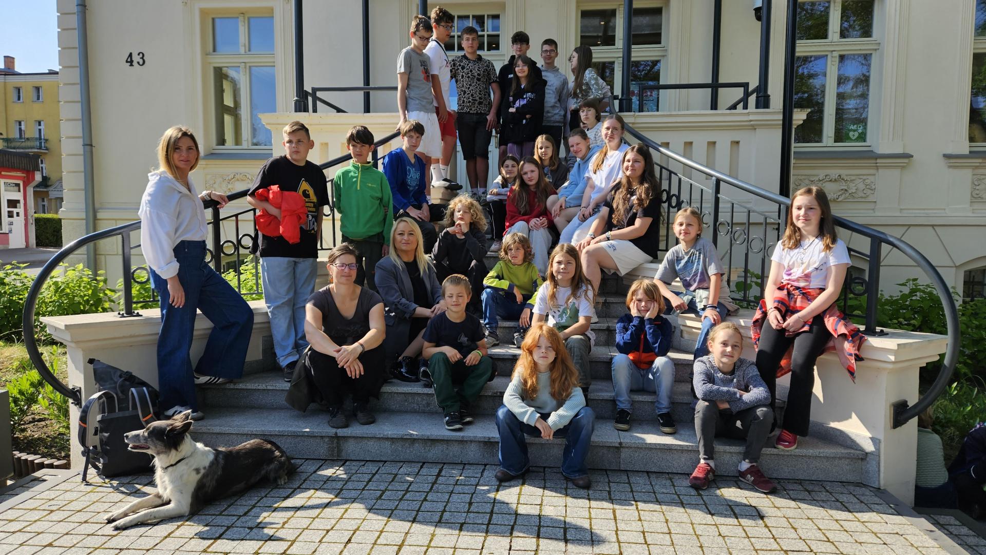 Uczniowie siedzą na schodach przed budynkiem w kolorze jasnym