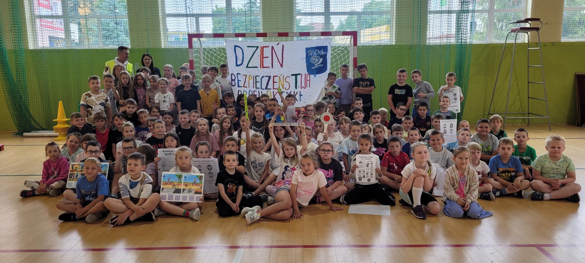 Duża grupa uczniów siedzi i stoi na sali. Między nimi widać baner  z napisem kolorowym Dzień Bezpieczeństwa