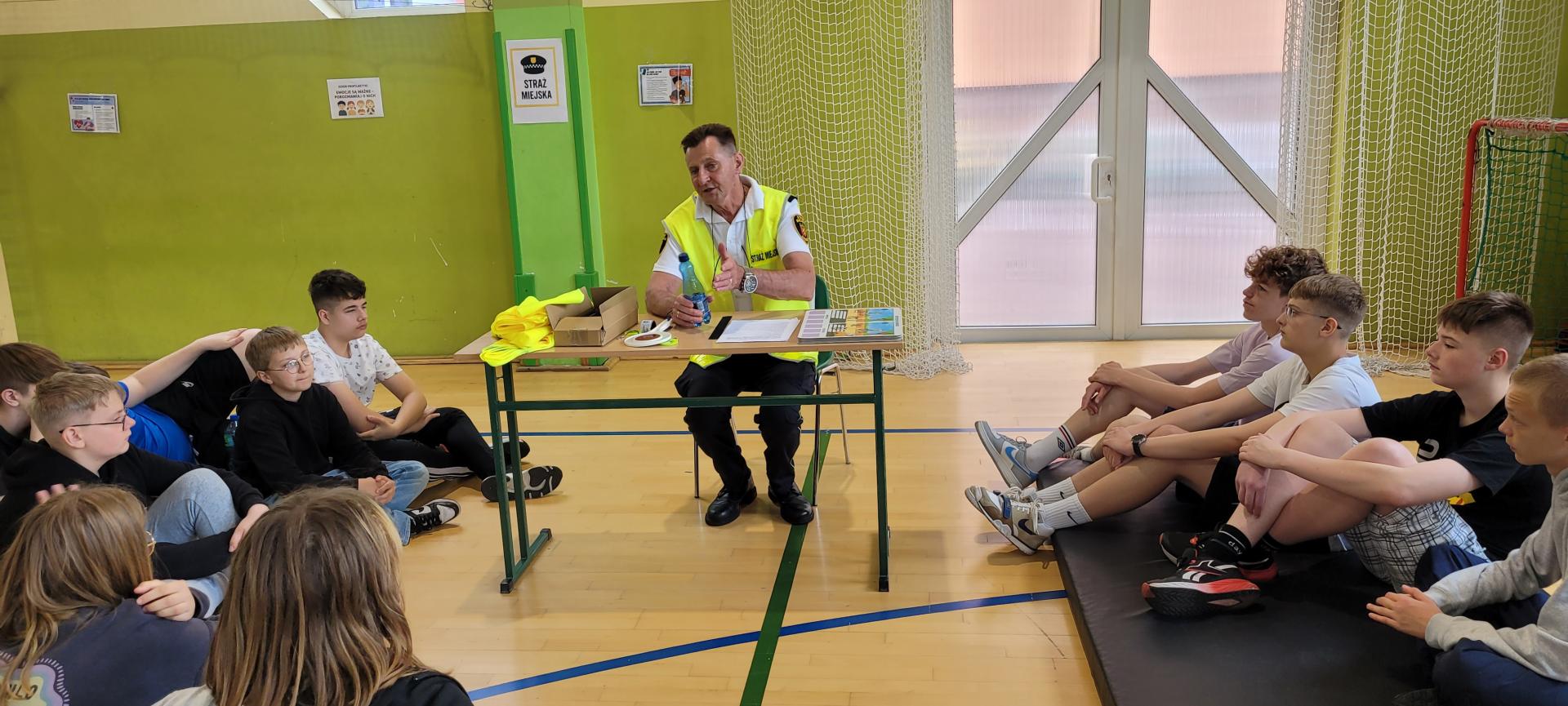 Centranie Komendant siedzi przy stoliku, po bokach uczniowie siedzą na materacach