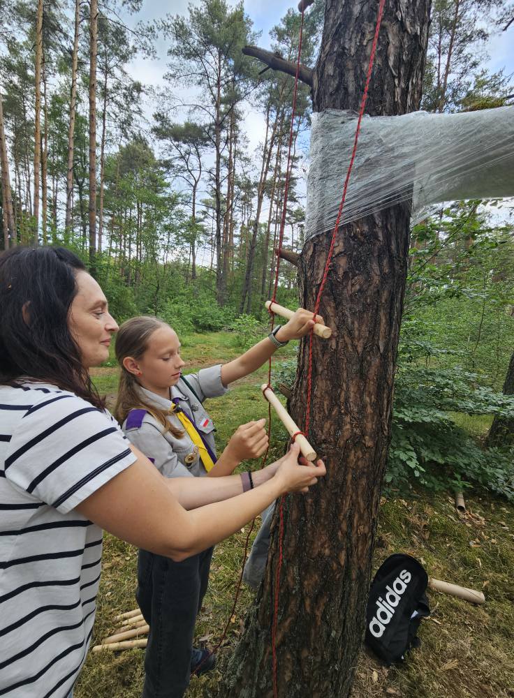 Kobieta wykonuje z dziewczynką w harcerskim stroju drabinkę z rurek i czerownego sznurka drabinka wisi na drzewie 