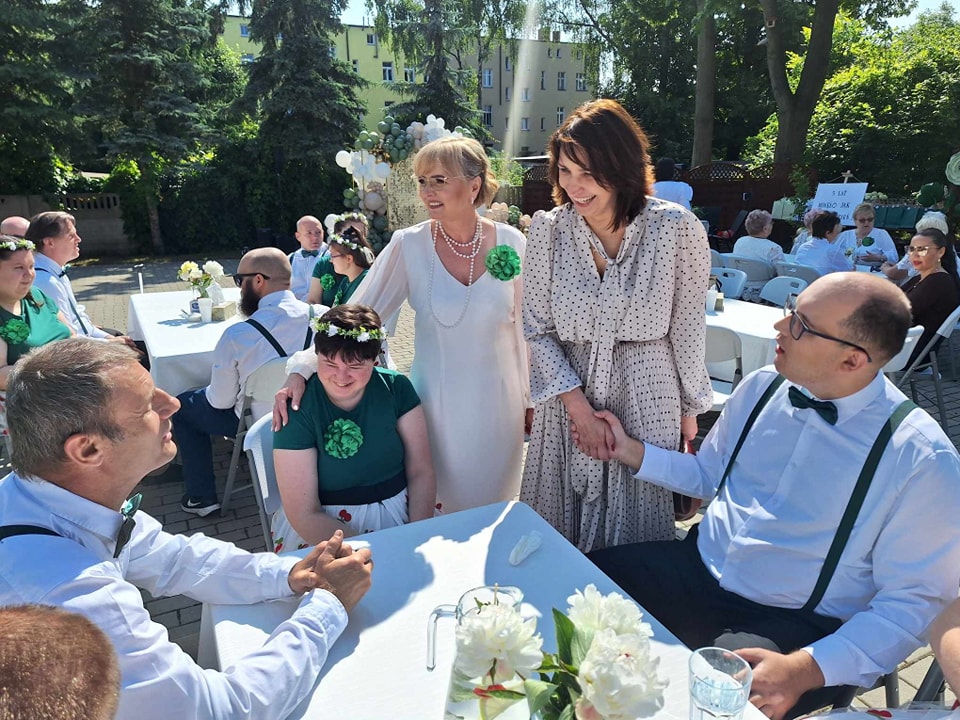 Po lewej stoi pani burmistrz podaje dłoń uczestnikowi, obok po lewej kierownik ŚDS i DDS, przy stole siedzą uczestnicy ŚDS